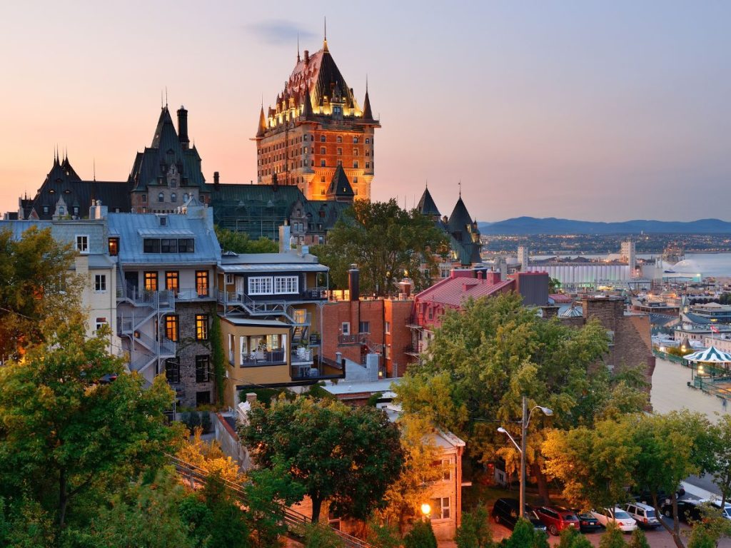Scorcio serale di Québec City con l'iconico e imponente Château Frontenac che svetta sopra gli edifici storici e gli alberi illuminati dalla luce calda del tramonto.