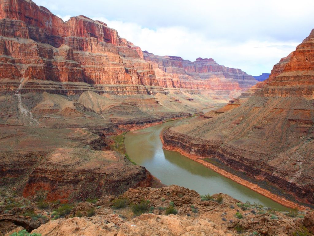 Veduta panoramica dall'alto del Grand Canyon con un fiume verde-marrone che scorre tra maestose pareti rocciose stratificate nei toni del rosso e dell'arancione.