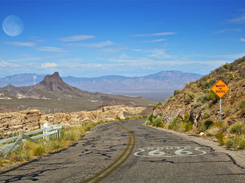 Strada asfaltata deserta con la scritta Route 66 impressa sulla carreggiata, circondata da paesaggi desertici e colline sotto un cielo azzurro chiaro.