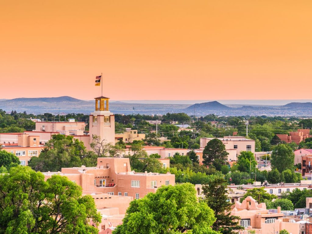Panorama cittadino di Santa Fe al tramonto con edifici in stile adobe color terracotta immersi nel verde e uno sfondo di montagne e cielo dalle sfumature calde.